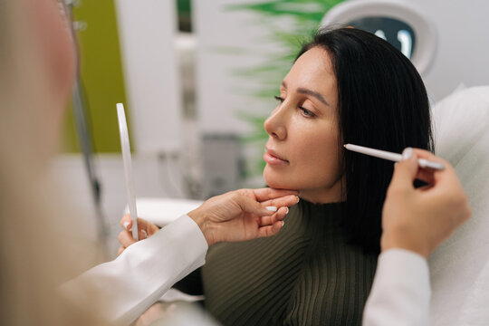 Side view of woman looking in mirror while cosmetologist marking face for aesthetic procedure, preparing for beauty treatment in cosmetology clinic. Concept of facial anti-aging, skincare treatment.