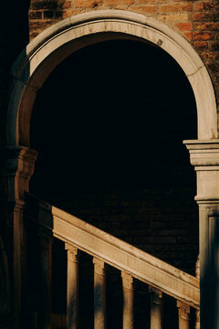 Architectural detail of the spiral staircase at Palazzo Contarini del Bovolo in Venice, with arched stone columns and side light creating long, dramatic shadows.