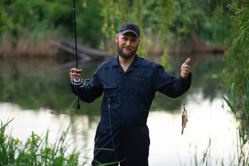 Young bearded fisherman holding a freshly caught perch on a spinning rod with a silicone lure, successful fishing on the river.