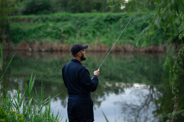 Young bearded man in a black cap standing by the river, fishing with a spinning rod, half-turned view, surrounded by lush green grass.