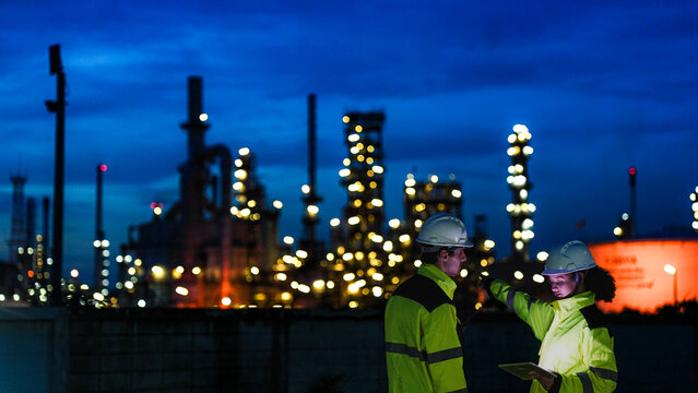 Petrochemical engineers in PPE conduct a night shift inspection at a refinery. They use a tablet for real-time data monitoring and auditing of the processing plant.