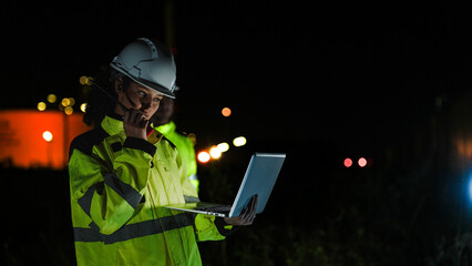 Two industrial workers in hard hats and high-vis gear work at night. They use a laptop and tablet...