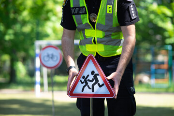 Ukrainian patrol police officer in a bright high-visibility vest with "Police" inscription holding a "Children" road sign on the street.