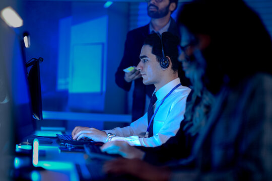 IT support team lead oversees a junior agent handling a complex technical issue. He offers real time advice while reviewing progress on screen in a busy operations center.