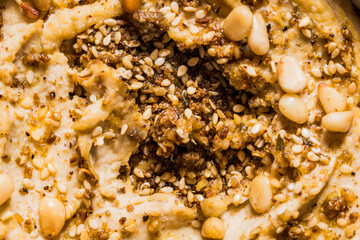 Close-up overhead view of smooth, golden hummus served in a white ceramic bowl. The creamy chickpea dip features a well in the center filled with olive oil, topped with toasted pine nuts, dark za'atar