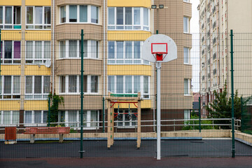 Basketball hoop in urban playground with residential building and fenced area, benches and greenery visible in the background. High quality photo
