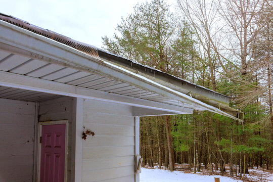 Snow rests on roof broken gutter while trees stand tall in winter morning.