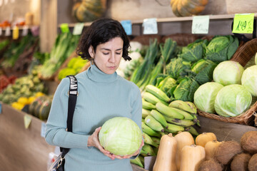 Fototapeta premium Woman pulls cabbage in his hands and chooses which one to cook soup and salad. Female customer came to a supermarket for fresh vegetables