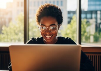 Young Black woman wearing glasses smiles while working on a laptop in a bright office setting