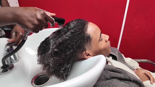 Right-side view of teen boy having his afro hair washed at a salon sink by a stylist