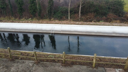 A drone shot showing a long, straight concrete water canal cutting through green agricultural fields. In the background, the Prealpine foothills of Lombardy under an overcast sky.  © marcobir