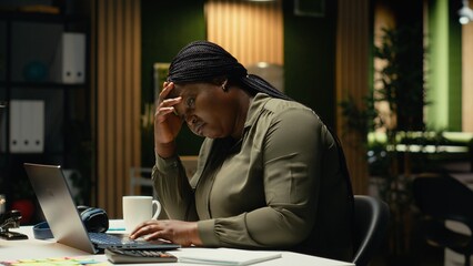 Obraz premium Black business employee realizing the night time alone in office, pushing limits despite clear signs of fatigue and work tension. Workplace challenges and sleep deprivation. Camera B.