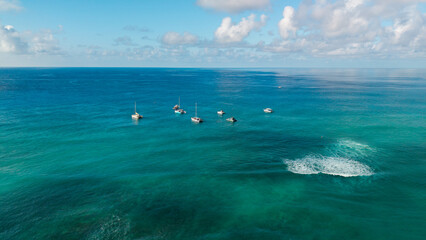 Aerial view of boats anchored in calm turquoise ocean water with a small wave breaking nearby, tropical seascape under a clear sky. © Bpato Media