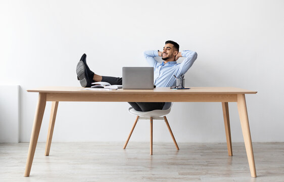 Relief After Finished Work. Smiling businessman relaxing on chair sitting at table resting feet on table, using pc, happy guy leaning back at workplace holding hands behind head, feeling pleased