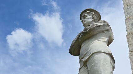 Obraz premium Maryborough, Australia - 4 January 2026. A carved stone soldier at the Maryborough War Memorial faces outward with arms crossed, set against a blue sky with scattered clouds.
