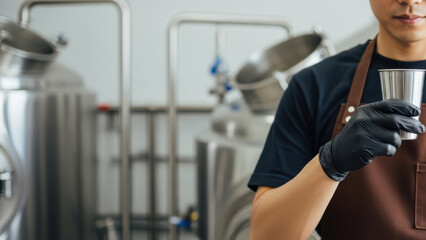 Fototapeta premium Focused brewery worker wearing brown apron and black gloves holding metal tasting cup in modern stainless steel craft beer brewing facility with fermentation tanks