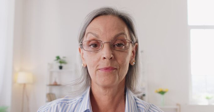 Webcam view headshot of a senior woman on a conference call. Old gray haired lady looks at the camera, puts on her glasses, and changes her expression from strict and serious to smiling and friendly