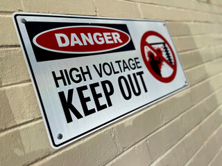 Naklejka premium Closeup of metal “Danger High Voltage Keep Out” safety sign on brick wall, selective focus emphasizing industrial electrical hazard warning.