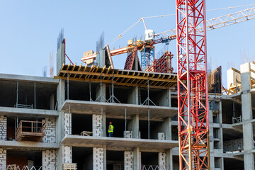 Construction worker wearing a safety vest examines the building structure with scaffolding and a crane visible on site, showcasing a multi-story construction project in an urban area
