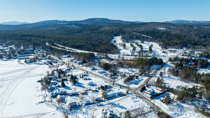 Snowy aerial view of Schroon Lake, NY with frozen lake, snowmobile tracks, forested hills, and winter homes.