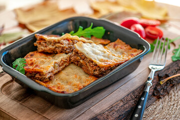 Fototapeta premium Lasagna placed in a black dish with salad and tomatoes on wooden table. Lasagna is served in a black dish on a wooden table with salad and tomatoes nearby.