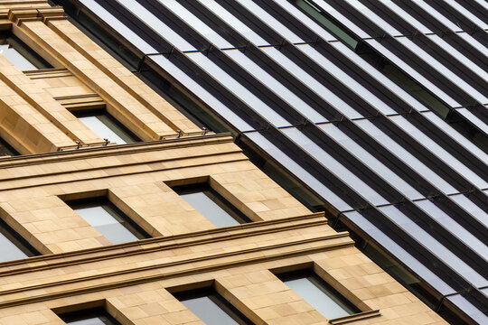 Close-up diagonal composition of heritage sandstone building meeting sleek modern glass facade, contrasting warm masonry details with cool reflective panels in urban cityscape.