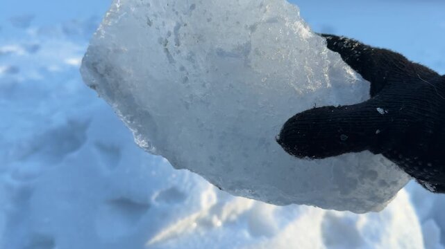 Hand Holding Flat Shard of Clear Ice in Winter. A gloved hand holds a large flat shard of clear ice against snow, sunlight highlighting its crystal-like structure and edges.