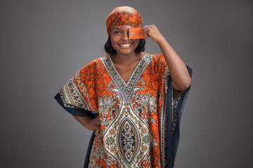 Obraz premium Studio portrait of a Nigerian woman wearing patterned traditional attire and headscarf, smiling while holding a card over one eye with her hand on her hip against a plain gray background.