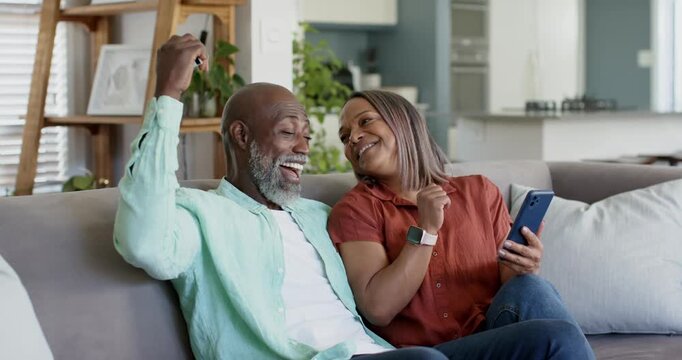 Senior couple sitting on couch, woman showing blue smartphone, man raising key fob celebrating
