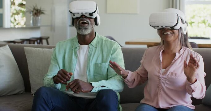 Diverse couple sitting on living room couch wearing white VR headsets, reacting to virtual stimuli