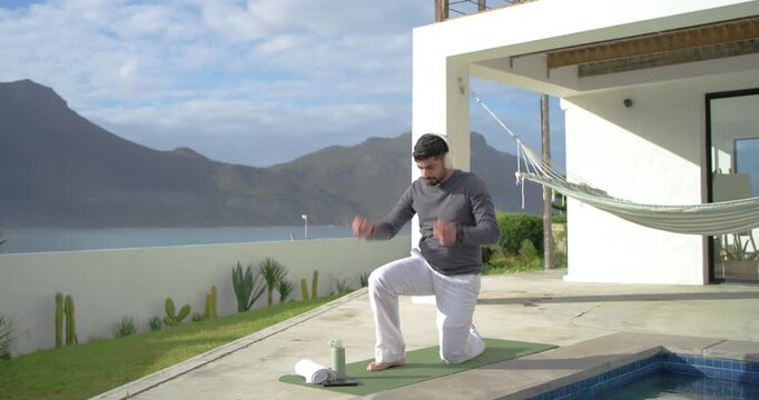 Indian man kneeling on green mat by pool, prepping workout, adjusting headset and drinking bottle