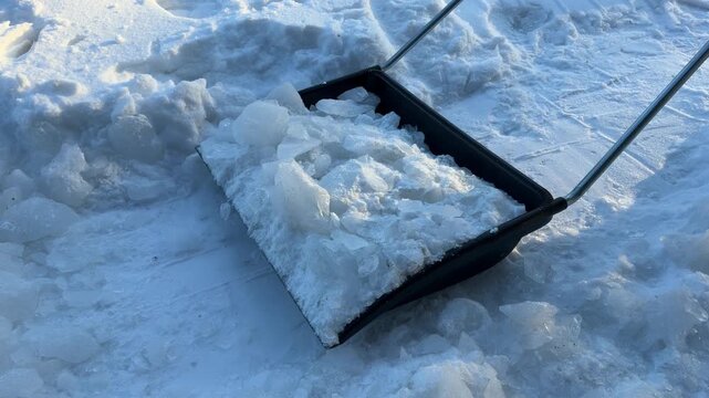 Pushing Ice Pile with Snow Shovel. A black snow shovel pushes a large pile of broken ice chunks across a snowy surface, clearing ice buildup in winter.