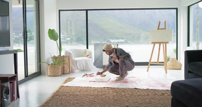 Indian man 20s crouching on studio floor canvas, painting roller, reloading tray as paint runs low