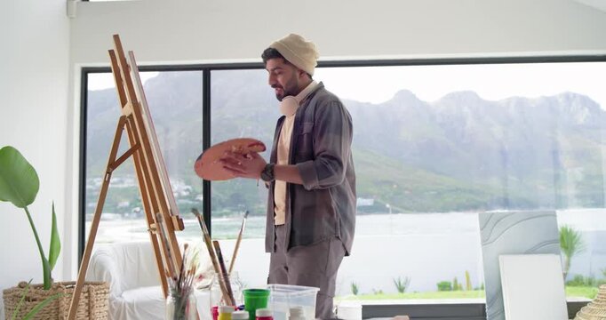 Indian male painter referencing view, dipping brush and painting canvas at home studio with palette