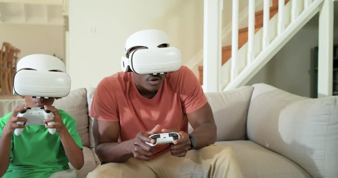 African American dad and son putting on white VR headsets, holding controllers on couch, playing VR