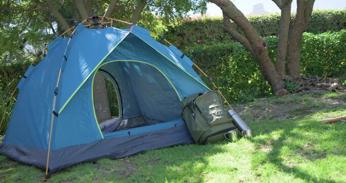 Camera is starting slow pan, revealing blue dome tent with backpack and thermos on shaded grass
