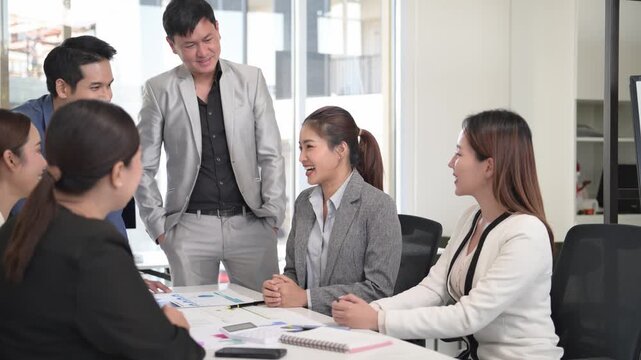 A group of people are sitting around a table in a conference room. There are five people in total, with two men and three women. They are all dressed in business attire, with one woman wearing a tie