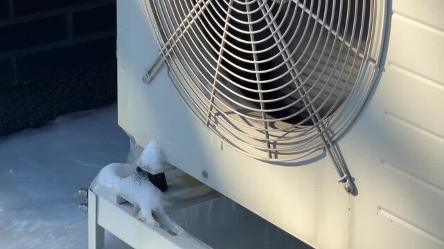 Close Up of Sunlit Heat Pump Fan and Grille. A close-up static shot shows a sunlit heat pump fan grille with ice accumulation at the bottom against a brick wall in winter.