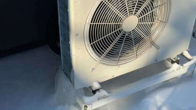 Heat Pump Outdoor Unit with Ice Buildup in Winter. A white heat pump stands in snow with ice at its base, the fan grille bright in winter sunlight, showing operation in freezing weather.