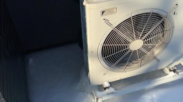 High Angle View of Heat Pump in Snow. A high-angle shot looks down at a snow-topped heat pump with ice at the base, sunlight illuminating the fan grille and white casing.