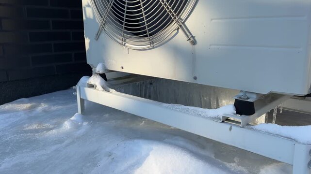 Frozen Base of a Heat Pump Unit in Winter. A close-up captures a white heat pump in snow with significant ice buildup at its base, the fan visible behind the grille.