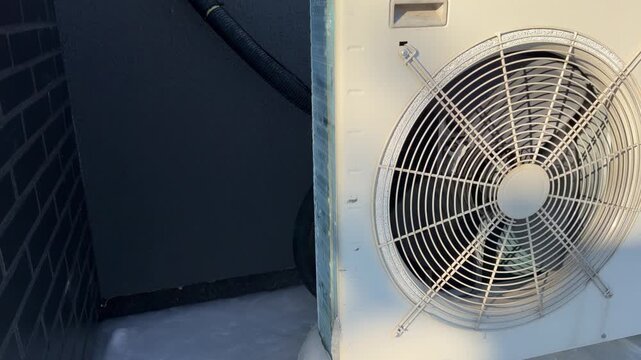 Side View of Heat Pump with Ice Accumulation. A side view shows a heat pump in snow with ice buildup on connection pipes and the bottom, set against a dark wall in winter.