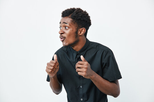 Young black man with dark skin wearing black shirt showing thumbs up gesture and looking sideways on white background. Positive expression and casual style with short hair.