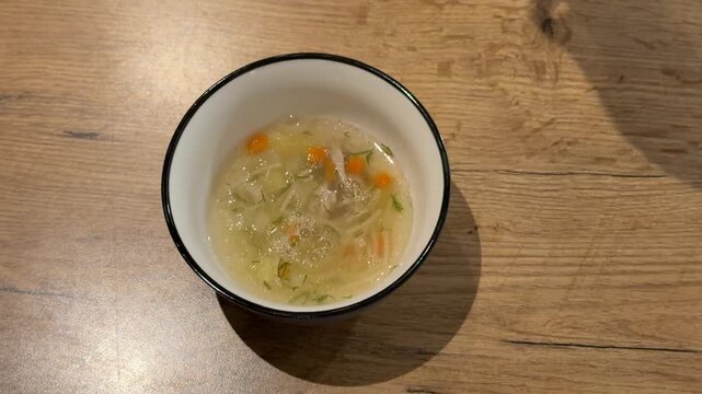 Pouring Vegetable Soup into a Bowl with a Ladle. A ladle pours hot vegetable soup with noodles and carrots into a white bowl on a wooden table, steam rising from the comforting dish.
