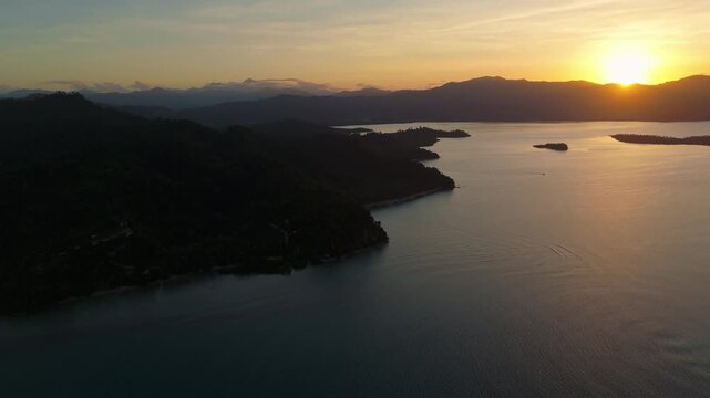 Cinematic aerial shot of the sun setting behind the tropical mountains of Port Barton. Vibrant golden sky reflecting on the calm sea at dusk in Palawan