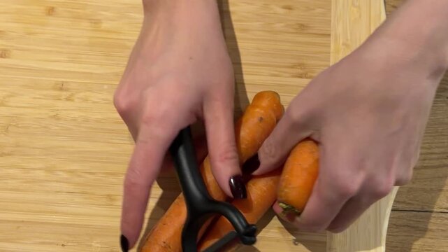 Preparing Carrots by Peeling Skin. A close-up shows hands peeling raw carrots on a wooden board with a black hand peeler, focusing on the repetitive cleaning motion.