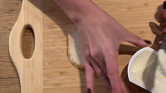Spreading Butter on a Slice of Toast Bread. A top-down shot shows hands spreading butter evenly over a slice of white toast on a wooden board, preparing a simple sandwich base.
