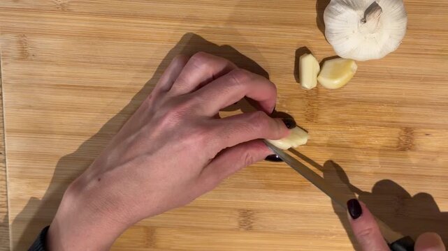 Dicing Garlic Cloves on a Wooden Board Top Down. A top-down shot displays hands dicing garlic cloves into tiny pieces on a wooden board, showcasing the precise preparation process.