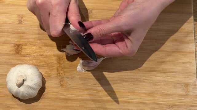 Hands Peeling Garlic Cloves on a Wooden Board. A close-up reveals hands using a small knife to peel skin from individual garlic cloves on a wooden board, preparing fresh ingredients.