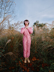 Obraz premium Outdoor scene with a person in a pink jumpsuit standing among tall grasses, hands in prayer, wearing a mask, autumnal setting, calm mysterious mood.
